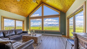 A spacious room with a high ceiling, large windows offering a view of the countryside, a leather sofa, and a wooden cabinet. - Mountain View Home w/Hot Tub - Close to Downtown! (Bozeman)