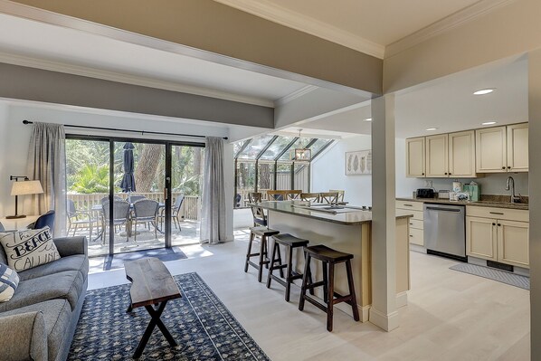 Amplia sala de estar con comedor, cocina con armarios de colores claros y puerta corrediza de vidrio que da a un balcón.