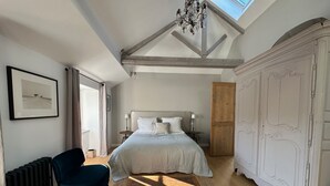A bedroom with a large bed, a wooden coffee table with a book, a blue chair, a radiator, and a white wardrobe. - Stylish two bedroom apartment with river views (Pont-Aven)
