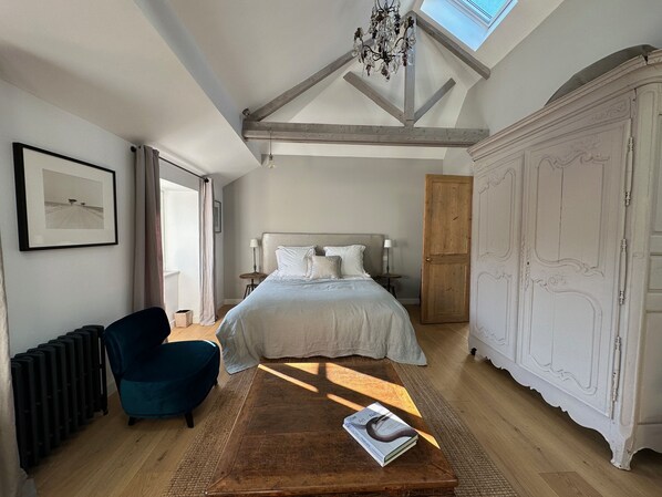 Un dormitorio con una cama grande, una mesita de madera con un libro, una silla azul, un radiador y un armario blanco.