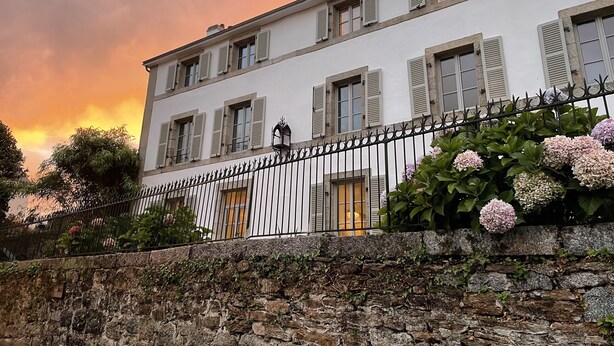 A white building with multiple windows and shutters, a stone wall in the foreground, and a metal fence. - Stylish two bedroom apartment with river views (Pont-Aven)