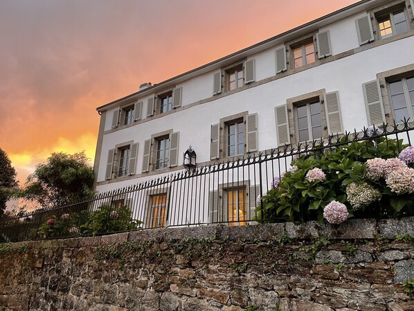 A white building with multiple windows and shutters, a stone wall in the foreground, and a metal fence.