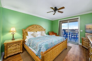 Quarto com cama, mesas de cabeceira de madeira, ventoinha no teto, varanda com cadeiras e televisão.