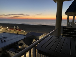 Uma varanda com vista para uma praia e um pôr do sol, um edifício com telhado vermelho, e uma escadaria que leva ao andar superior.