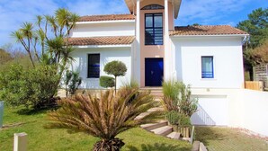 Une maison à deux étages avec une entrée centrale, un petit jardin devant et un ciel d’un bleu éclatant.