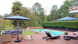 Une terrasse en bois, aménagée avec du mobilier d’extérieur, notamment un canapé, des fauteuils et des parasols, donnant sur une piscine.