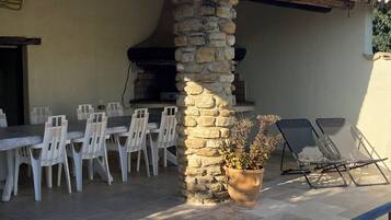 Une terrasse extérieure avec des piliers en pierre, un barbecue, des chaises en plastique blanches et un pot de fleurs.
