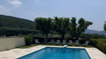 Une piscine rectangulaire à l’eau d’un bleu limpide, entourée d’une terrasse carrelée. Plusieurs chaises longues sont alignées le long du bord de la piscine, et une rangée d’arbres et d’arbustes se trouve au fond.