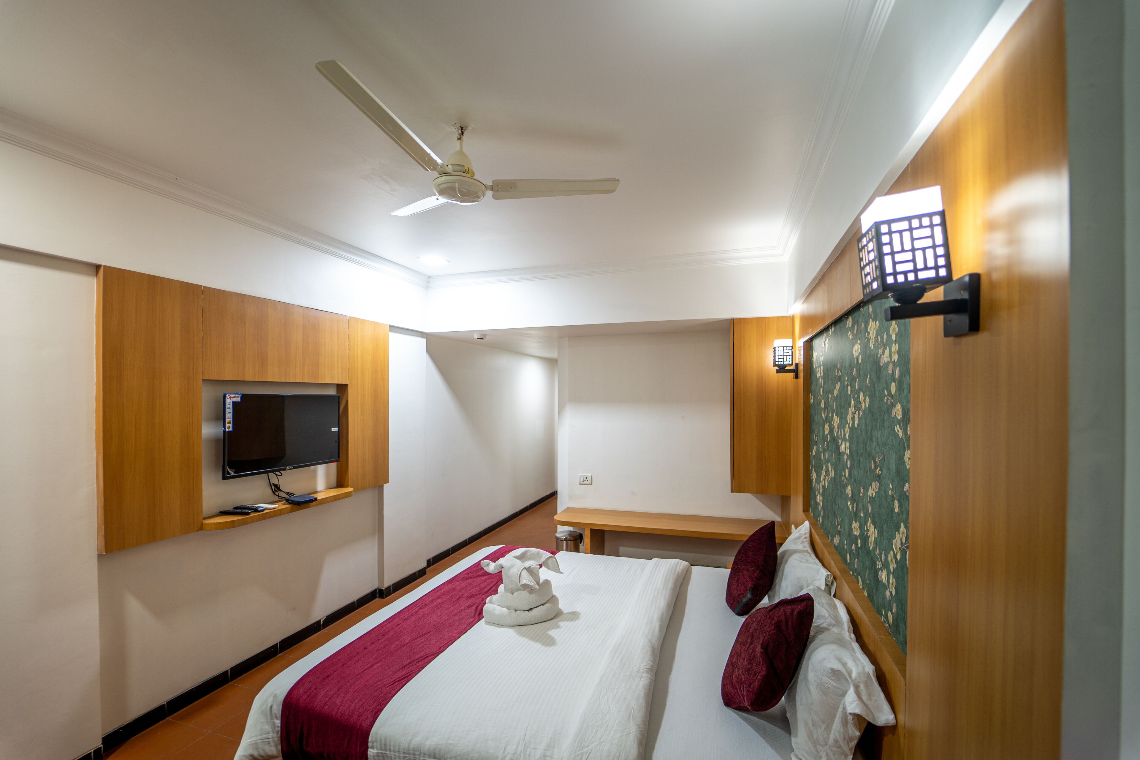 A hotel room with a bed, a flat-screen TV mounted on the wall, a ceiling fan, and a small table with a lamp.