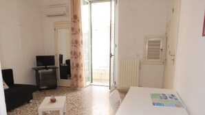 Ein Zimmer mit gefliestem Boden, einem weißen Tisch mit einer Karte, einem schwarzen Sofa, einem kleinen weißen Tisch mit einem dekorativen Gegenstand und einem Fernseher auf einem Schrank.