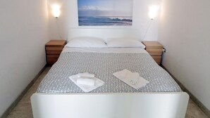 Ein ordentlich bezogenes Bett mit einer gemusterten Tagesdecke, zwei Kissen und einem gefalteten Handtuch. Darüber hängt ein Bild von einer Strandansicht.