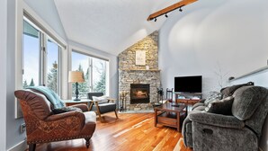 A living room with a stone fireplace, wooden flooring, and a large window offering a view of trees. - Rock Pond Condo 45 - Beautiful Ski in/Ski out condo unit on Saddleback! (Rangeley)