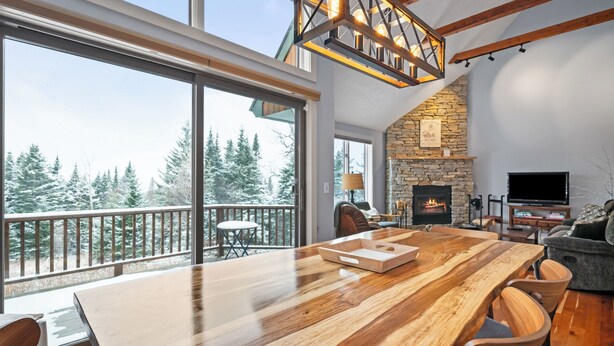 A spacious living area with a large wooden table, stone fireplace, and a balcony with a view of trees. - Rock Pond Condo 45 - Beautiful Ski in/Ski out condo unit on Saddleback! (Rangeley)