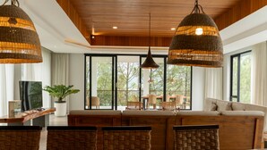 A spacious living area with a large wooden table, wicker chairs, and a sofa. There are large windows with a view of trees outside. - Atsuka Hill View 8BR Vila with Private Heated Pool (Cimenyan)