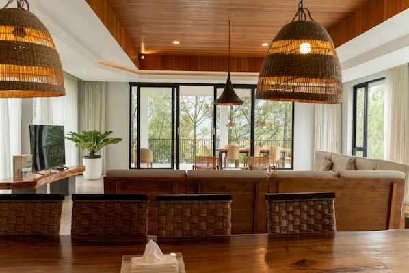 A spacious living area with a large wooden table, wicker chairs, and a sofa. There are large windows with a view of trees outside.