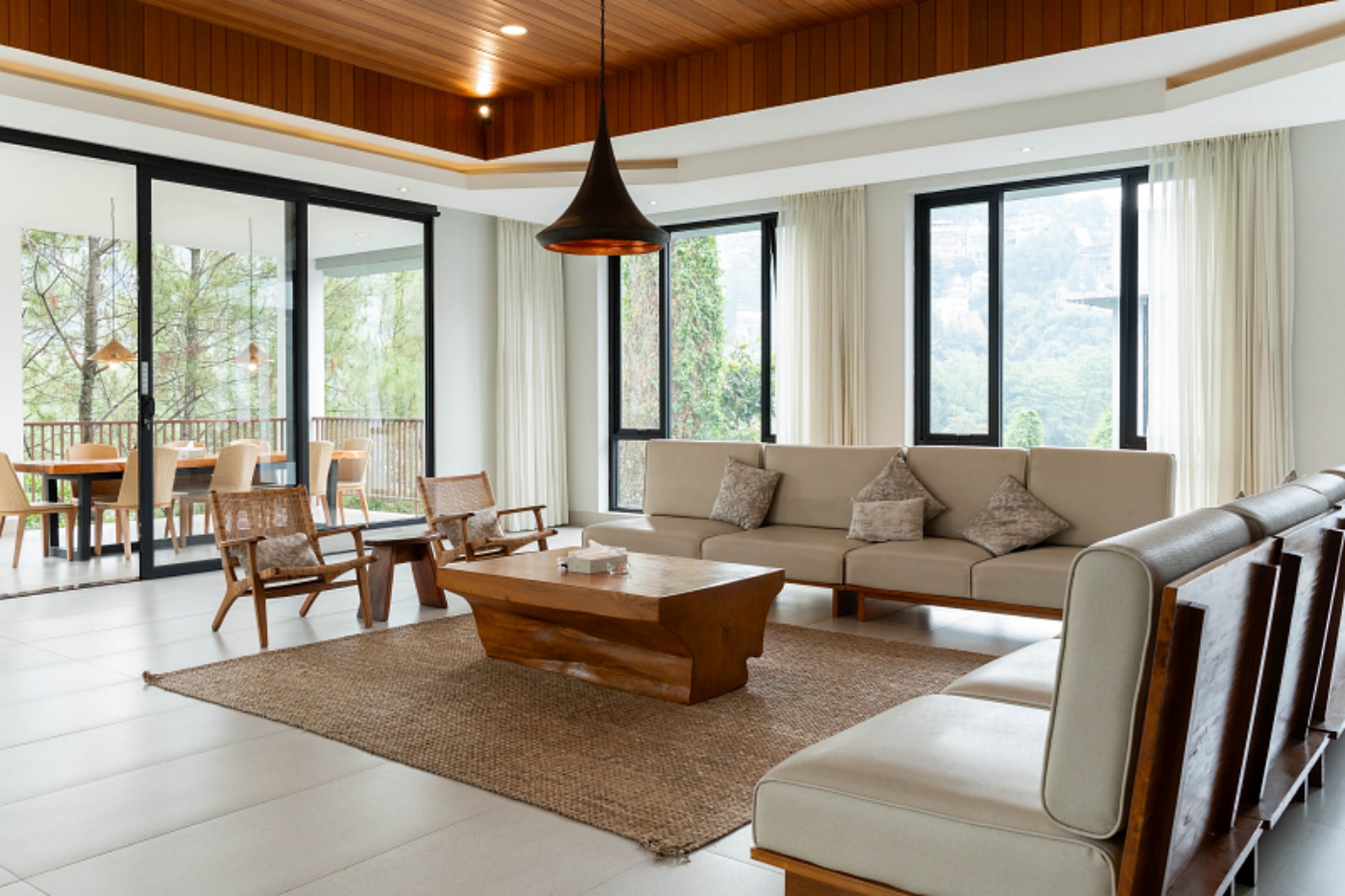 A spacious living room with a sofa, coffee table, and chairs, large windows with curtains, and a wooden ceiling.