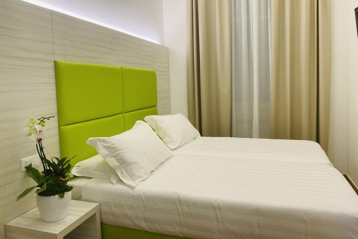 Habitación de hotel con cabecera verde, ropa de cama blanca, una planta en la mesita de noche y cortinas color beige.