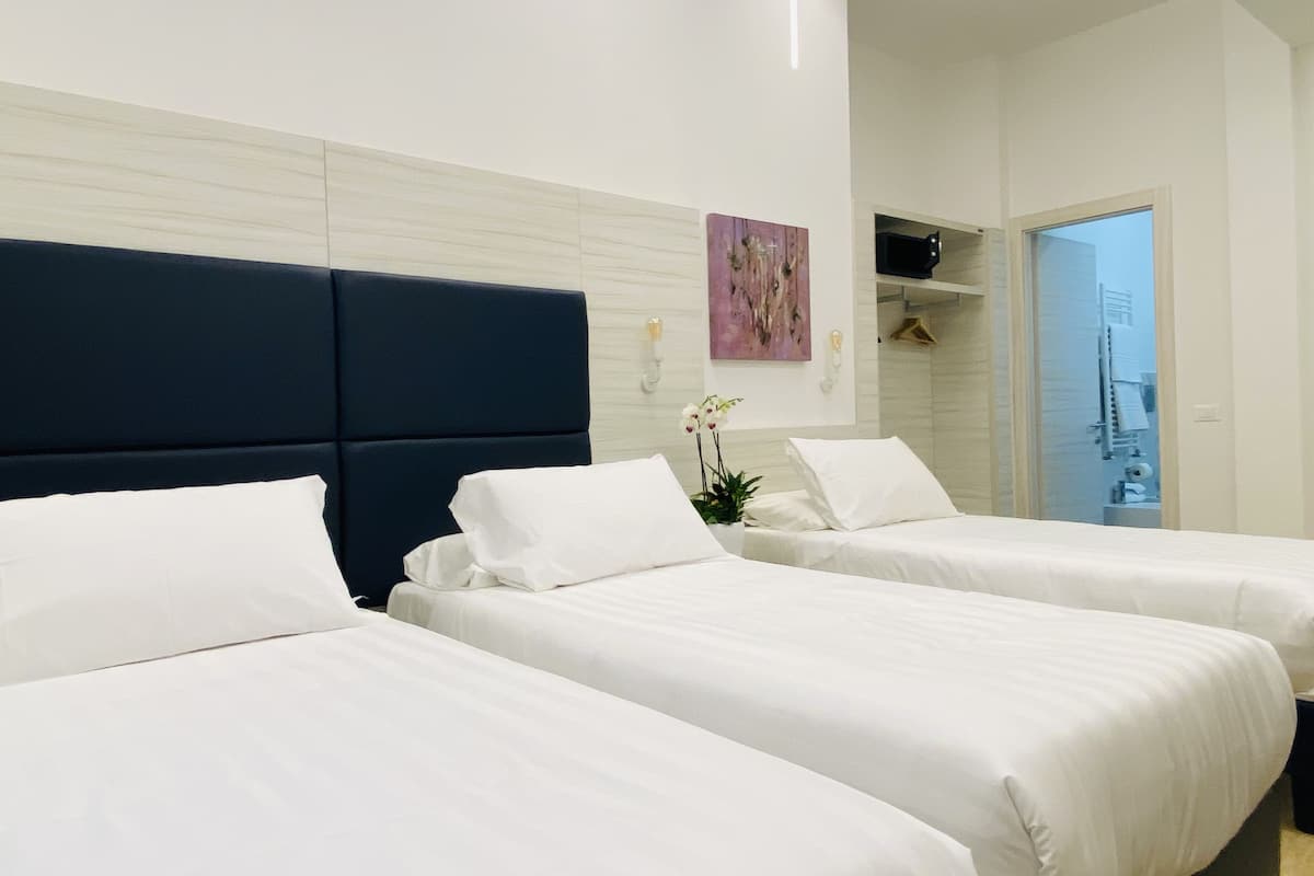 Habitación de hotel con tres camas individuales, cabecera oscura, pared blanca con un cuadro y vista a un balcón.