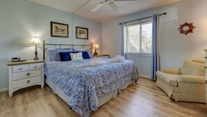 Ein Schlafzimmer mit Bett, Nachttischen, Deckenventilator, einem Fenster mit Vorhängen und einem Sessel.