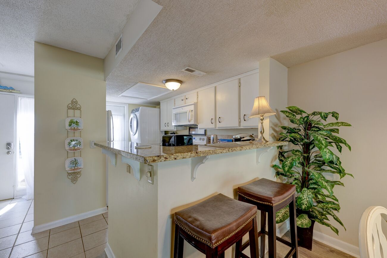 A kitchen with white cabinets, a microwave, and a countertop. There are two stools and a potted plant.