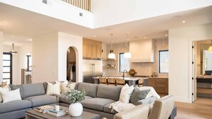 A spacious living room with a large gray sectional sofa, a coffee table with books and a vase, and a kitchen in the background with wooden cabinets and stainless steel appliances. - Golden Hour: Desert Color Lagoon + Beaches (St. George)