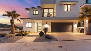 A modern two-story house with a garage, a balcony with outdoor furniture, and a well-lit entrance. - Golden Hour: Desert Color Lagoon + Beaches (St. George)