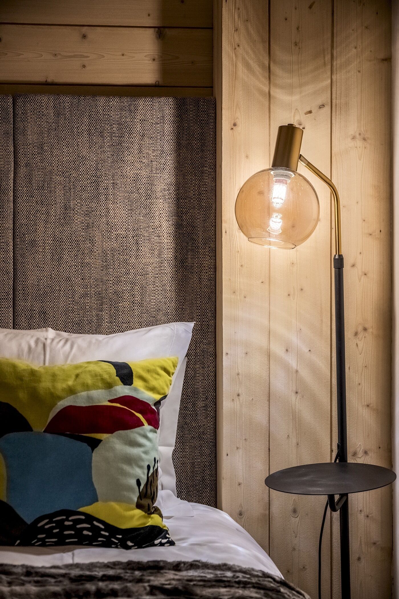 Un lit avec un oreiller coloré et une lampe de sol moderne dotée d’un abat-jour transparent.
