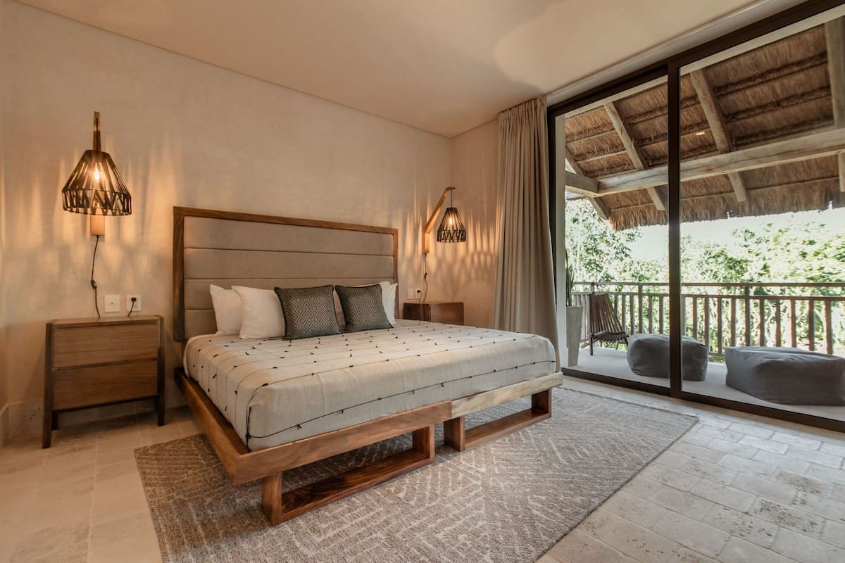 Une chambre avec un grand lit, une tête de lit en bois, deux tables de chevet et une petite table de nuit. Elle dispose d’un balcon doté d’un toit de chaume et de mobilier d’extérieur.