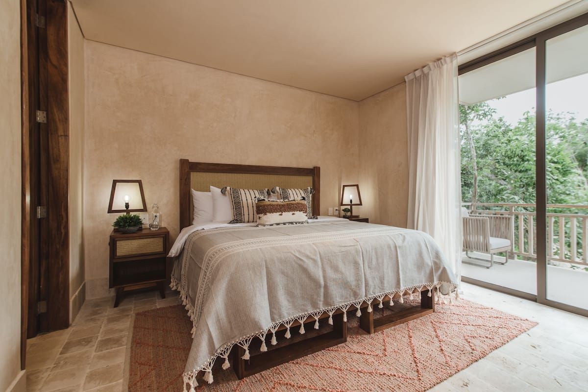 Une chambre dotée d’un grand lit, de deux tables de chevet avec des lampes, d’une vue sur l’extérieur grâce à une porte-fenêtre coulissante et d’un balcon aménagé avec du mobilier d’extérieur.