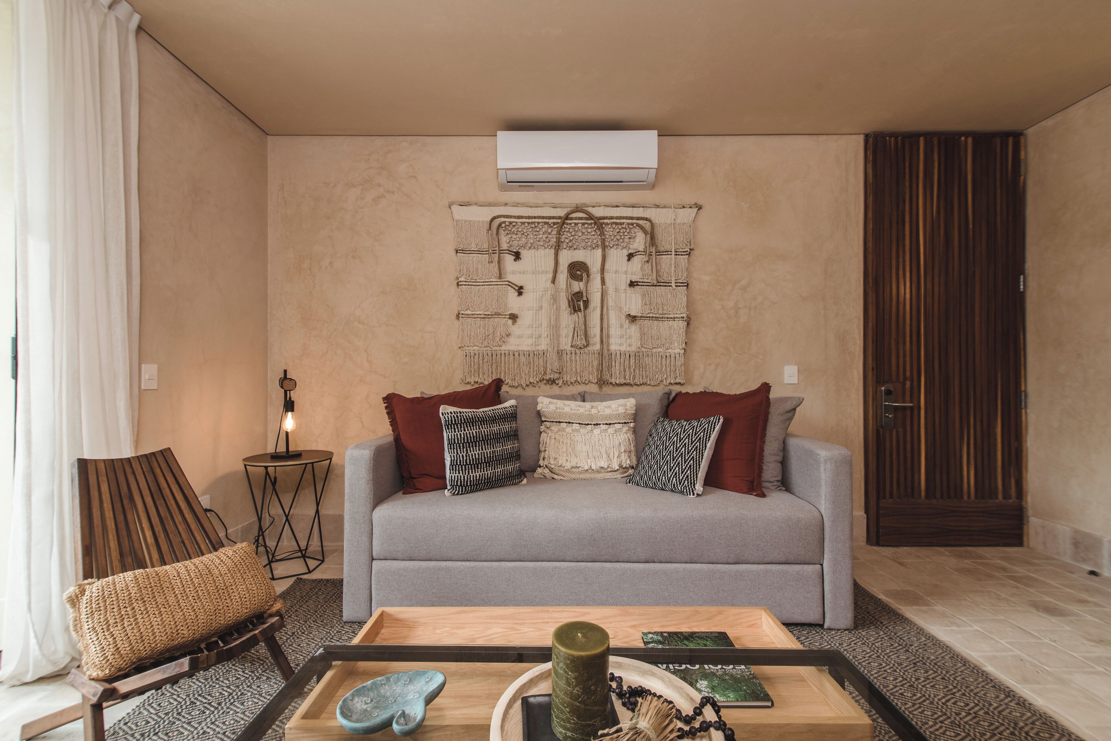 Ein Wohnzimmer mit grauem Sofa, gemusterten Kissen, einem Holztisch mit dekorativen Gegenständen, einer Wand-Klimaanlage und einem Wandteppich.
