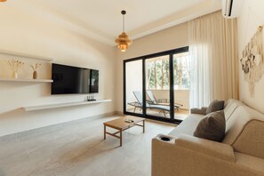 A modern living room with a flat-screen TV, a wooden coffee table, a beige sofa, and a large sliding glass door leading to an outdoor area with a chair and a table. - Lumina at LOSANTOS Tulum (Tulum)