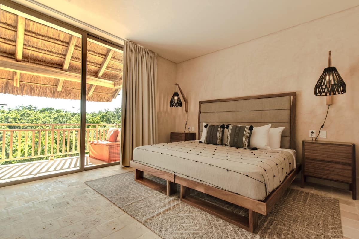 Une chambre avec un grand lit, une tête de lit en bois, une table de chevet, une lampe et une vue sur la verdure grâce à une porte-fenêtre coulissante.