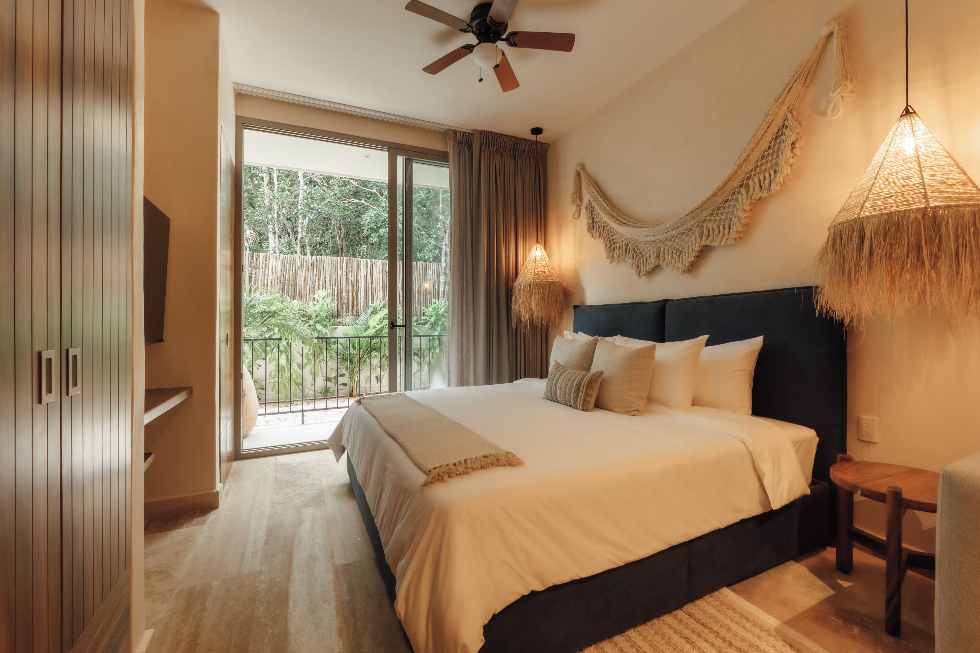 Een slaapkamer met een groot bed, een plafondventilator, een balkon met uitzicht op groen en een geweven lamp.