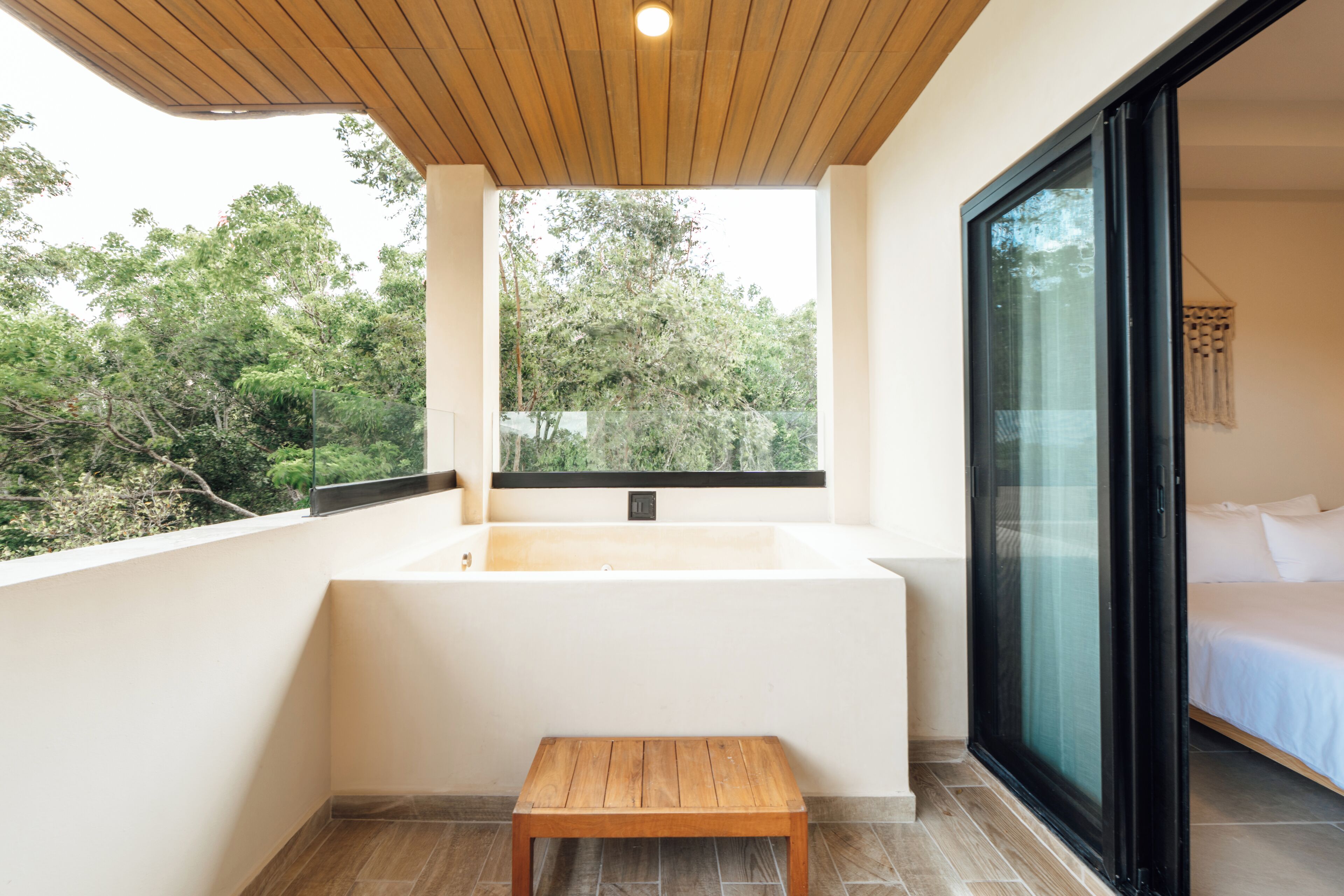 A modern bathroom with a bathtub, a wooden bench, and a large window offering a view of trees.