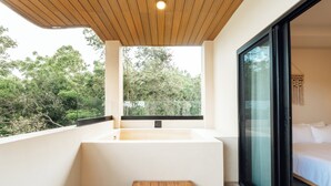 A modern bathroom with a bathtub, a wooden bench, and a large window offering a view of trees.