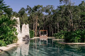 A serene outdoor pool area with a wooden deck and a wooden swing chair, surrounded by lush greenery and trees.