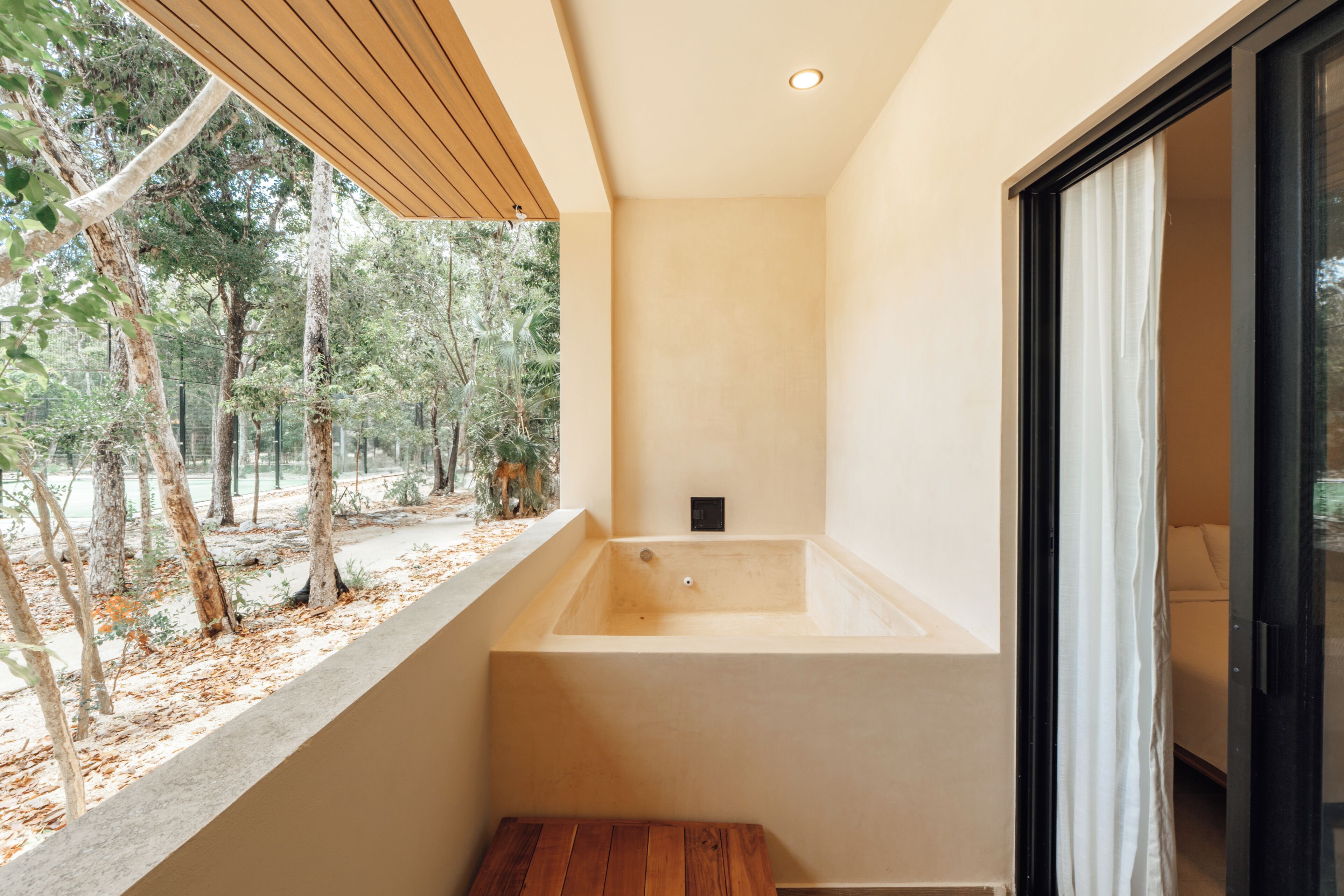 A modern bathroom with a large bathtub, a wooden bench, and a view of a forest.