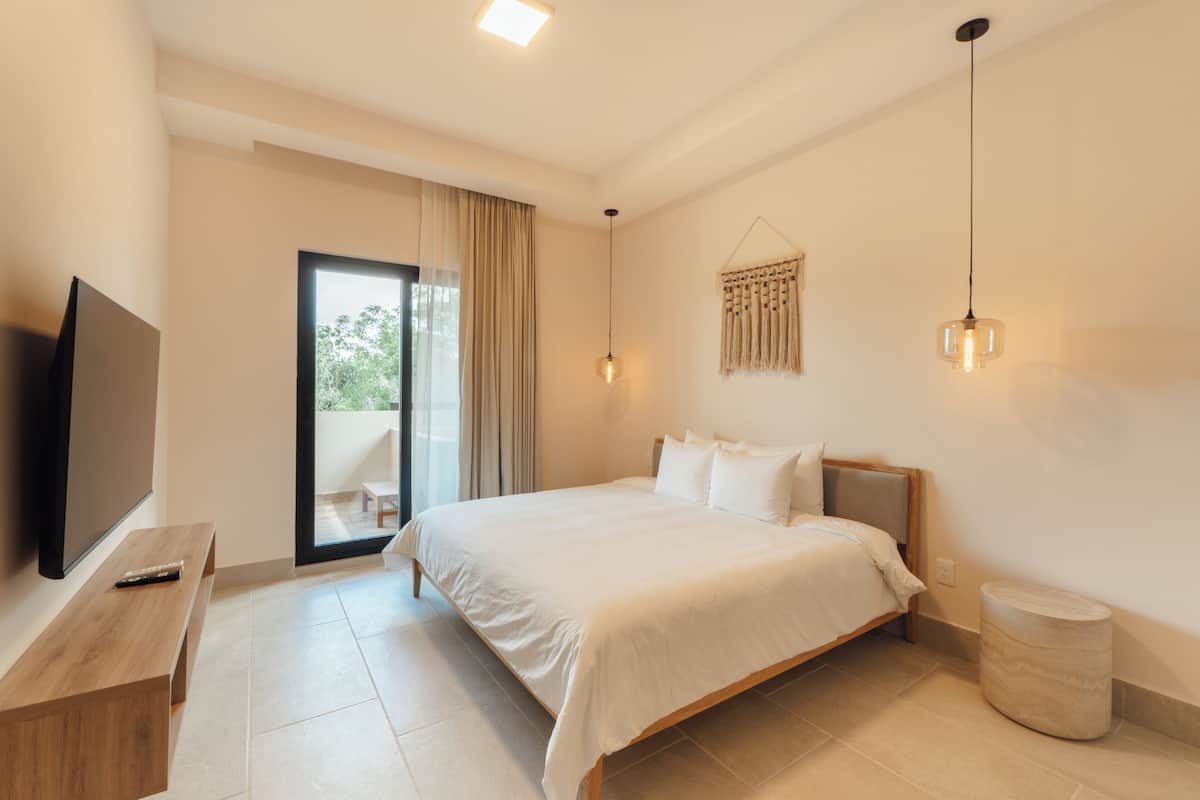 Una habitación de hotel moderna con una cama grande, un televisor sobre un mueble de madera, una mesita redonda y un balcón con cortinas.
