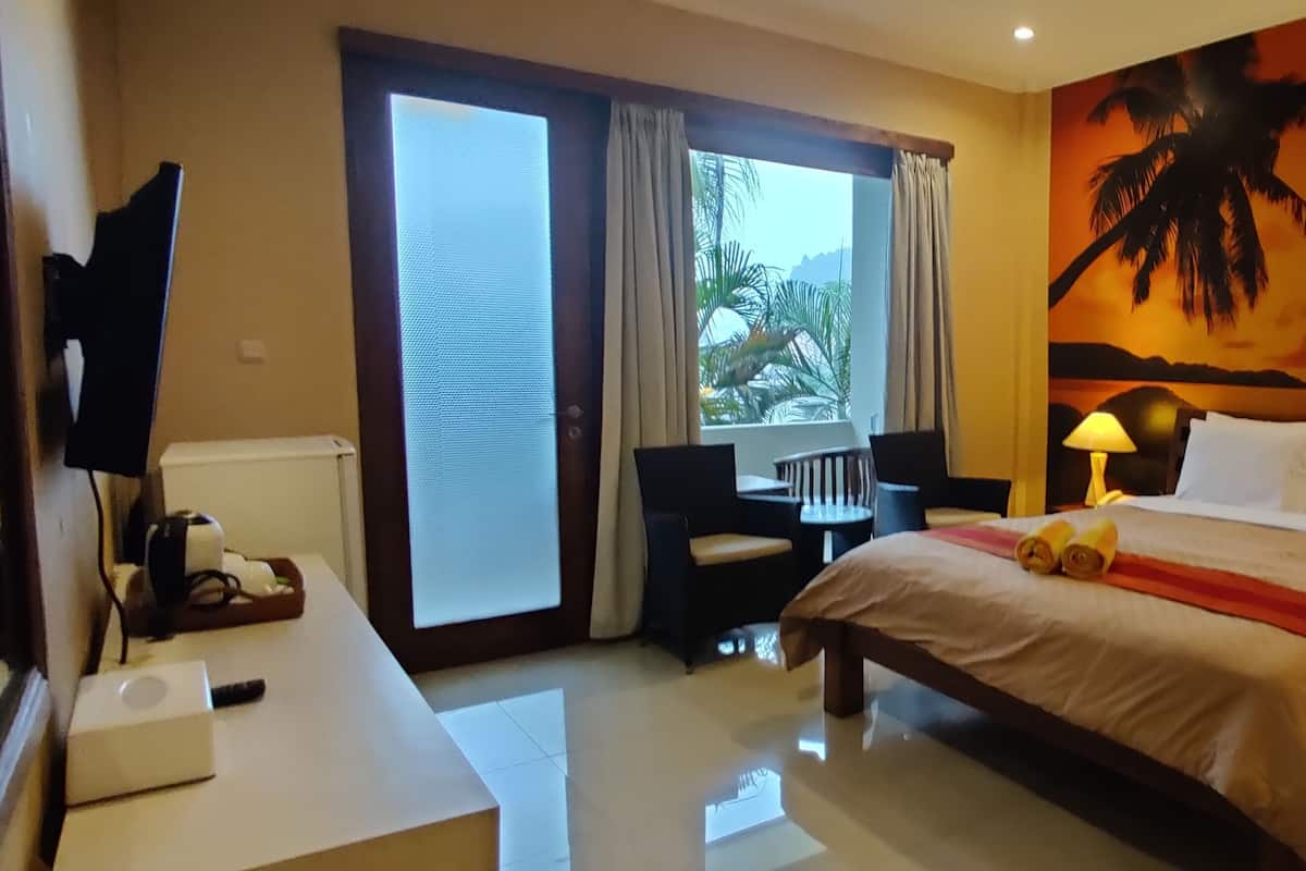 Una habitación de hotel con cama, escritorio, televisor, ventana con cortinas y un mural con un atardecer en la pared.