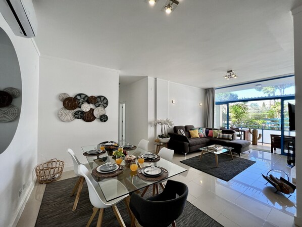 A modern living room with a glass dining table set for four, a black sofa, and a large window offering a view of the outdoors.