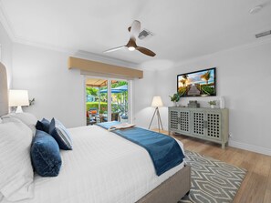 A bedroom with a bed, a ceiling fan, a TV, and a view of an outdoor area.