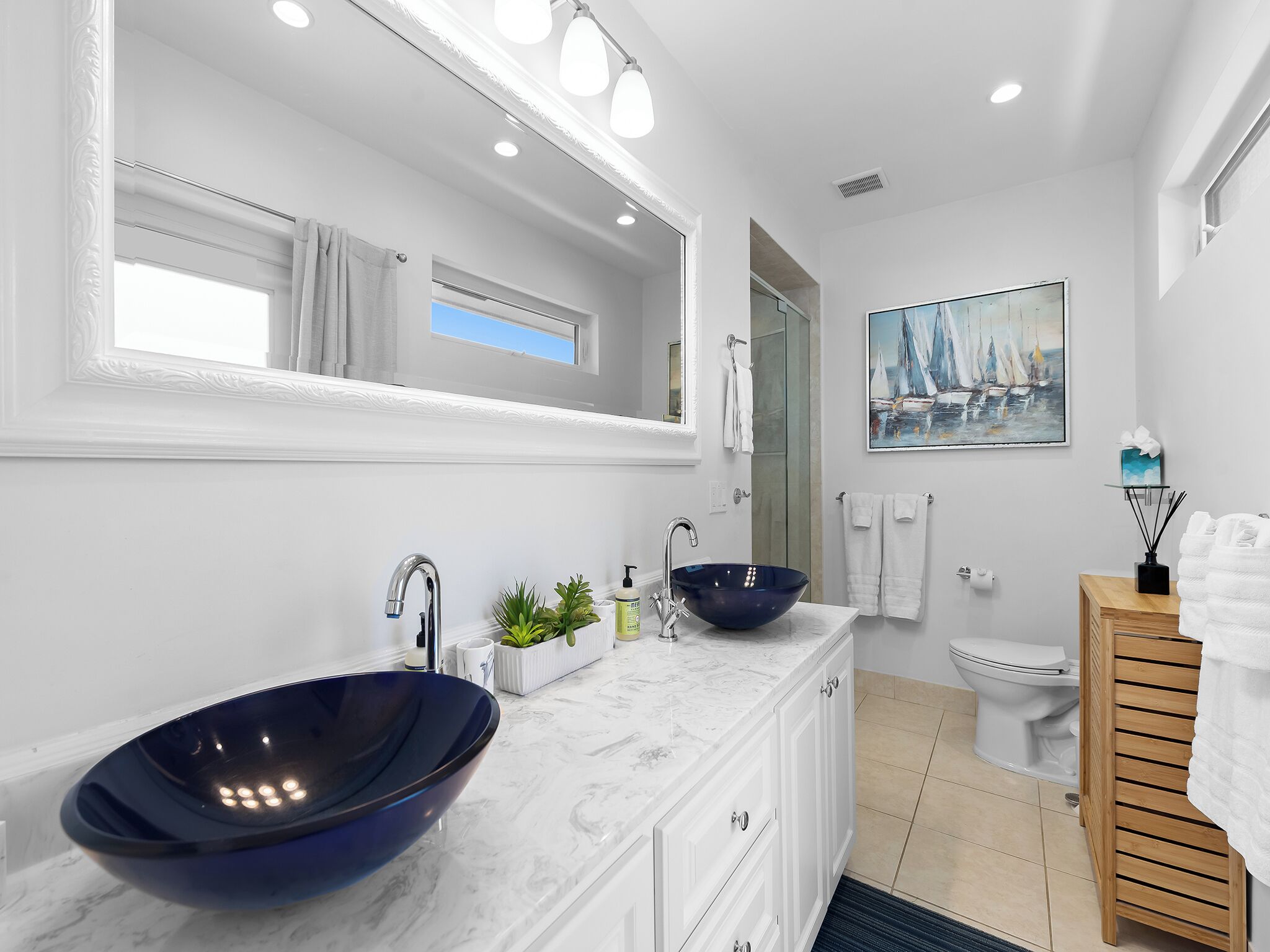 A modern bathroom with a large mirror, two vessel sinks, a wooden vanity, and a painting of sailboats.