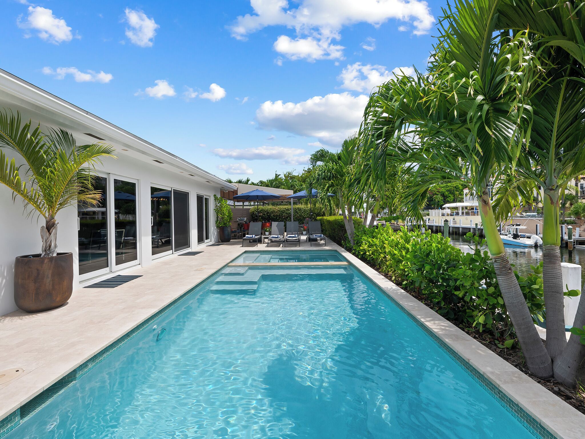A swimming pool with lounge chairs and umbrellas, surrounded by greenery and a building with large windows.