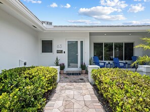 A modern house with a white exterior, a glass door entrance, and a patio area with blue chairs and potted plants.