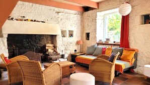 Una habitación acogedora con chimenea de piedra, muebles de mimbre, un sofá con cojines coloridos y una ventana con cortina roja.