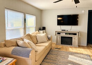 A living room with a beige sofa, a flat-screen TV mounted above a fireplace, and a window with blinds. - 1740 Ibis 2 Bedroom 2 Bathroom Home - Cozy Cottage (Surfside Beach)