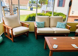 A covered outdoor seating area with two chairs and a sofa, a wooden table, and a view of a residential street. - 1740 Ibis 2 Bedroom 2 Bathroom Home - Cozy Cottage (Surfside Beach)