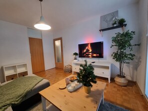 A cozy living room with a flat-screen TV displaying a fireplace, a coffee table with a Christmas tree, and a large potted plant.