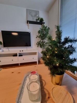 A modern living room with a flat-screen TV, white furniture, a small artificial Christmas tree, and a tray with candles.