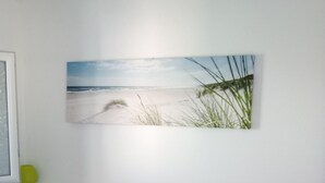 Une chambre à coucher avec un lit, une lampe fixée au mur, une table de chevet et un tableau représentant une scène de plage.